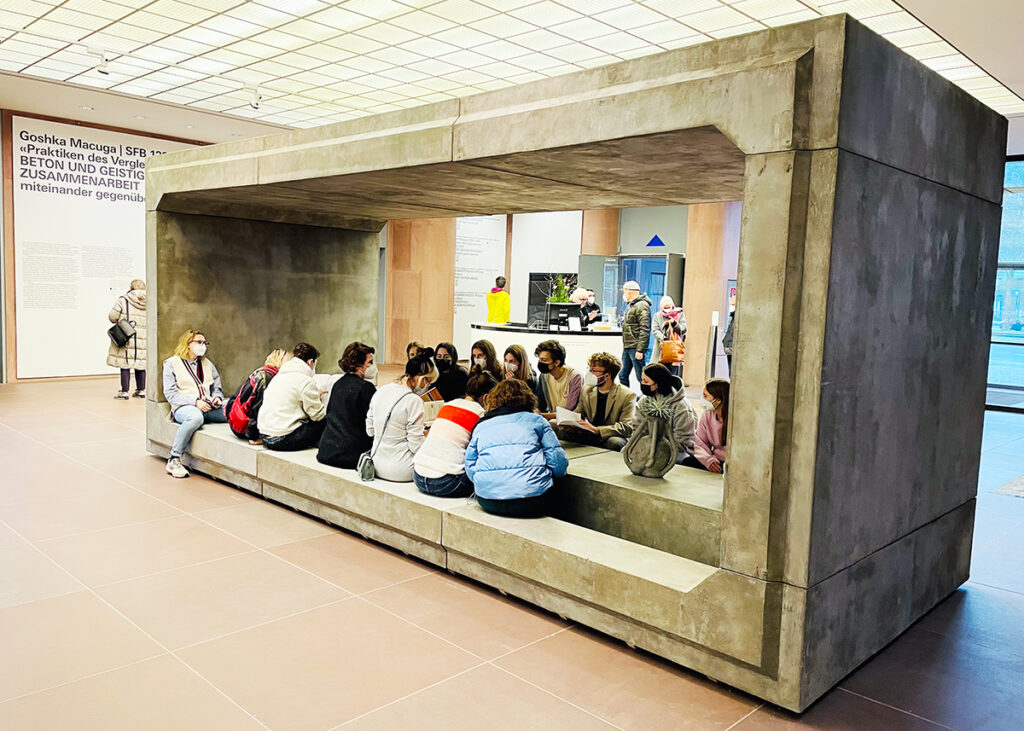 Beton-Quader in einem Foyer, in dem eine Gruppe von Menschen Platz genommen hat.