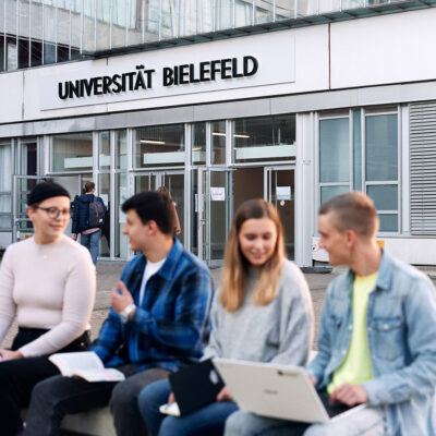 Vier Studierende sitzen vor dem Haupteingang der Universität Bielefeld und unterhalten sich.