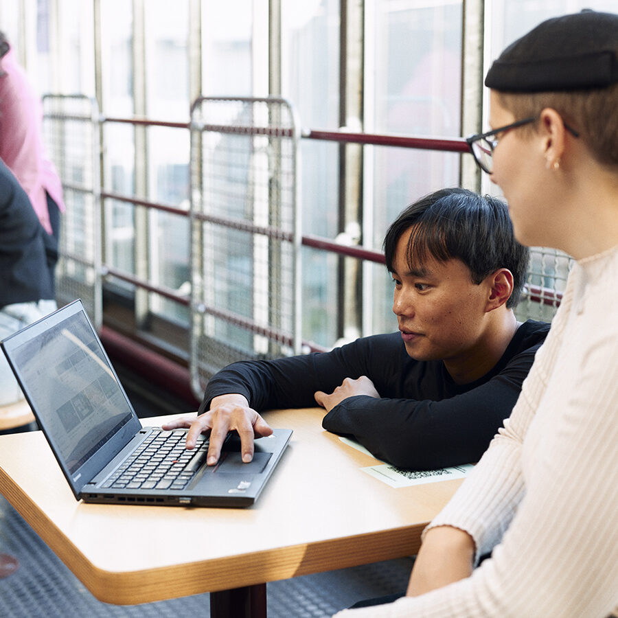 Zwei Studierende sitzen an einem Arbeitsplatz in der Uni und schauen gemeinsam auf ein Laptop.