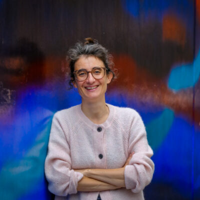 Die Philosophie-Professorin Christine Bratu lehnt lächelnd an einer bunten Wand, ihre Arme sind verschränkt.
