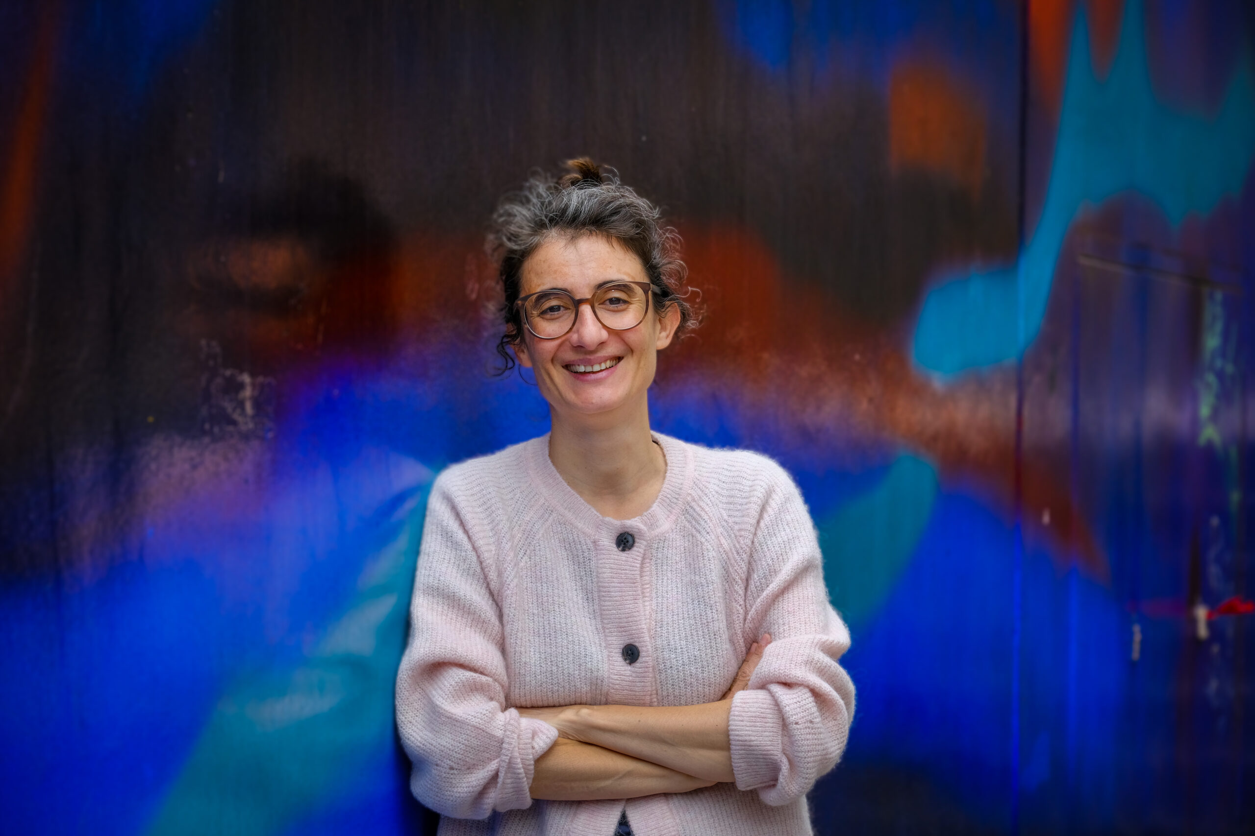 Die Philosophie-Professorin Christine Bratu lehnt lächelnd an einer bunten Wand, ihre Arme sind verschränkt.