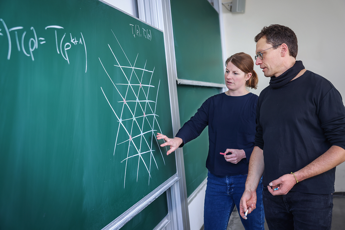 Two people stand at a blackboard with a recorded grid structure and discuss