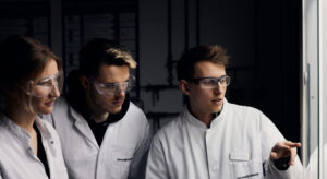 Drei Personen in weißen Laborkitteln und Schutzbrillen.