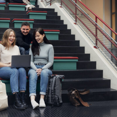 Drei Studierende sitzen mit einem Laptop auf der Treppe ins Nichts in der Universitätshalle.