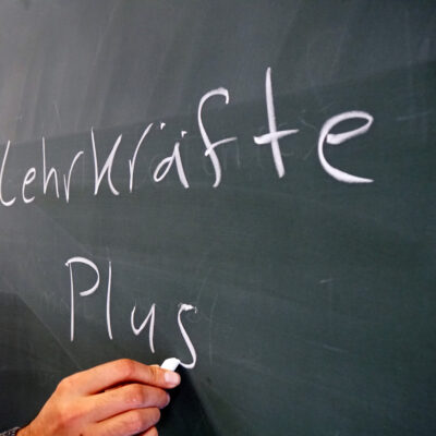 Hand schreibt mit Kreide “Lehrkräfte Plus” auf die Tafel