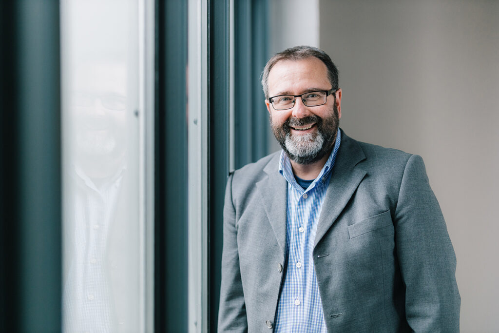 Professor Dr. Oliver Krüger steht rechts von einem Fenster. Er trägt ein graues Sakko.