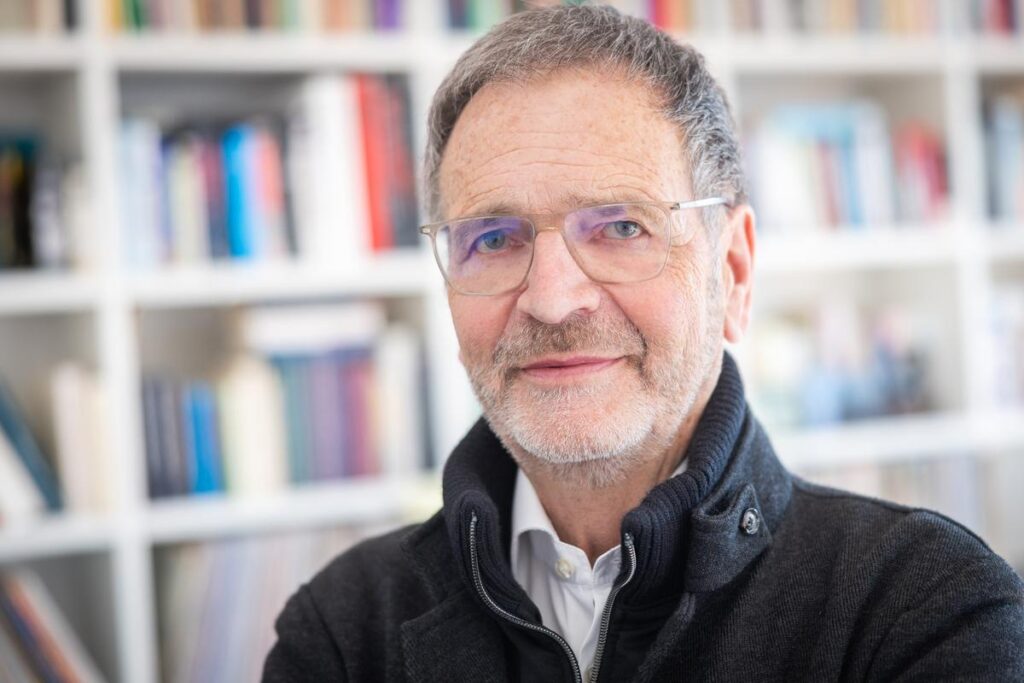 Professor Dr. med. Alexander Krämer, mit Brille und schwarzer Jacke, steht vor einem Bücherregal.