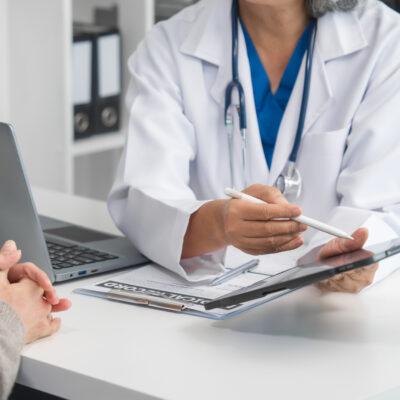 Ärztin spricht mit Patientin am Schreibtisch und hält dabei ein Tablet.