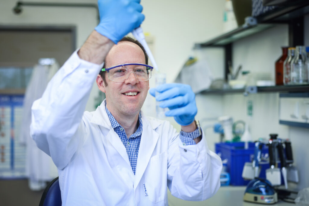 Professor Dr Joseph Hoffmann in a laboratory, wearing a white lab coat, blue gloves and safety goggles. He is holding a laboratory pipette and filling a liquid into a glass container. In the background, various laboratory equipment as well as glass flasks and vials can be seen on shelves.