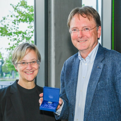 Rektorin Prof. Dr. Angelika Epple, links im Bild und Prof. Dr. Fred Becker, rechts im Bild hält die Ehrennadel der Universität Bielefeld.