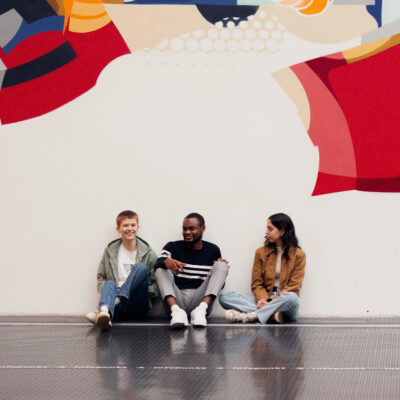 Drei Studierende sitzen auf dem Boden der Uni-Halle und unterhalten sich miteinander.