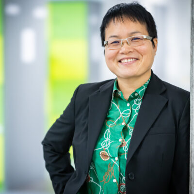 Professorin Dr. Minh Nguyen, in grünem Hemd und schwarzer Jacke, steht neben einer Säule mit Plakaten.