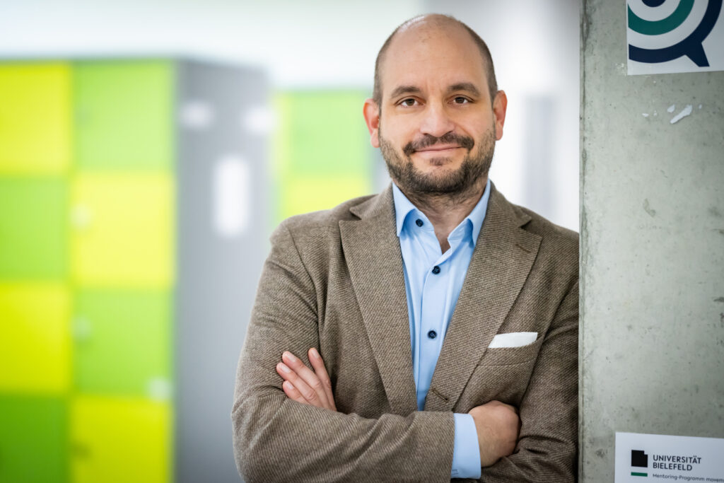 Professor Dr. Carsten Sauer, gekleidet in einem Anzug, steht neben einer Betonsäule, im Hintergrund grüne Schließfachschränke.