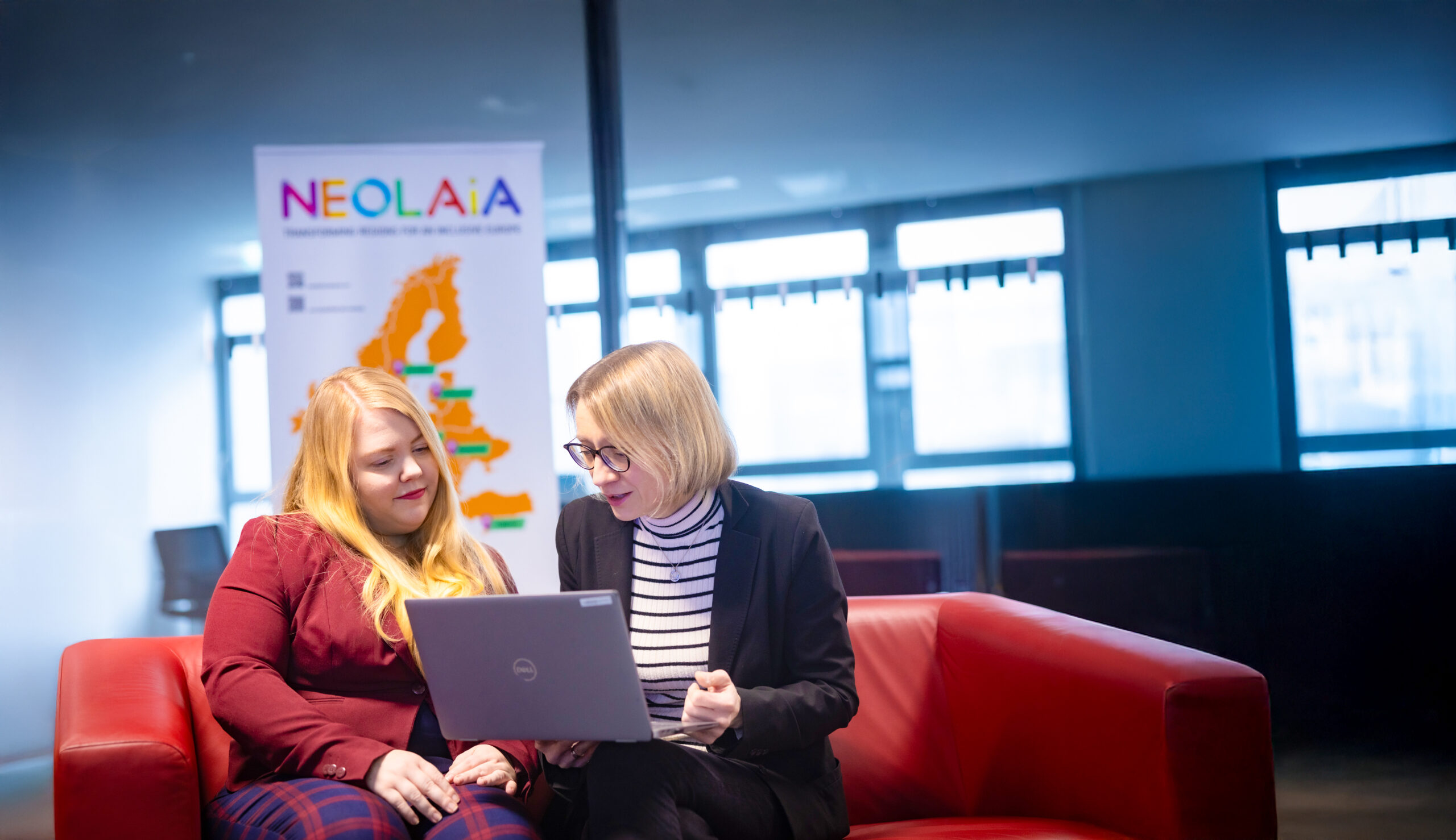 Amelie Labusch and Michaela Vogt are sitting on a couch ,looking at a laptop. Behind them is a NEOLAiA flag.