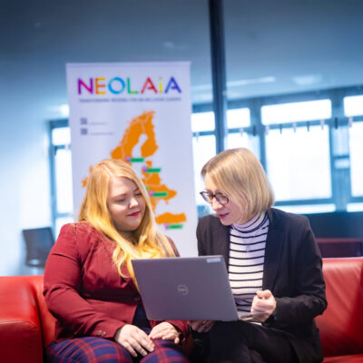 Amelie Labusch and Michaela Vogt are sitting on a couch ,looking at a laptop. Behind them is a NEOLAiA flag.