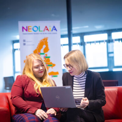 Amelie Labusch und Michaela Vogt sitzen auf einer Couch und schauen auf ein Laptop, hinter ihnen steht eine NEOLAiA Flagge.