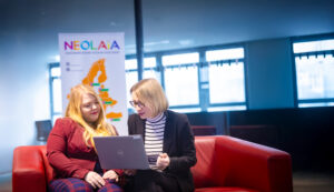 Amelie Labusch und Michaela Vogt sitzen auf einer Couch und schauen auf ein Laptop, hinter ihnen steht eine NEOLAiA Flagge.