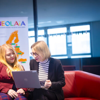 Amelie Labusch und Michaela Vogt sitzen auf einer Couch und schauen auf ein Laptop, hinter ihnen steht eine NEOLAiA Flagge.
