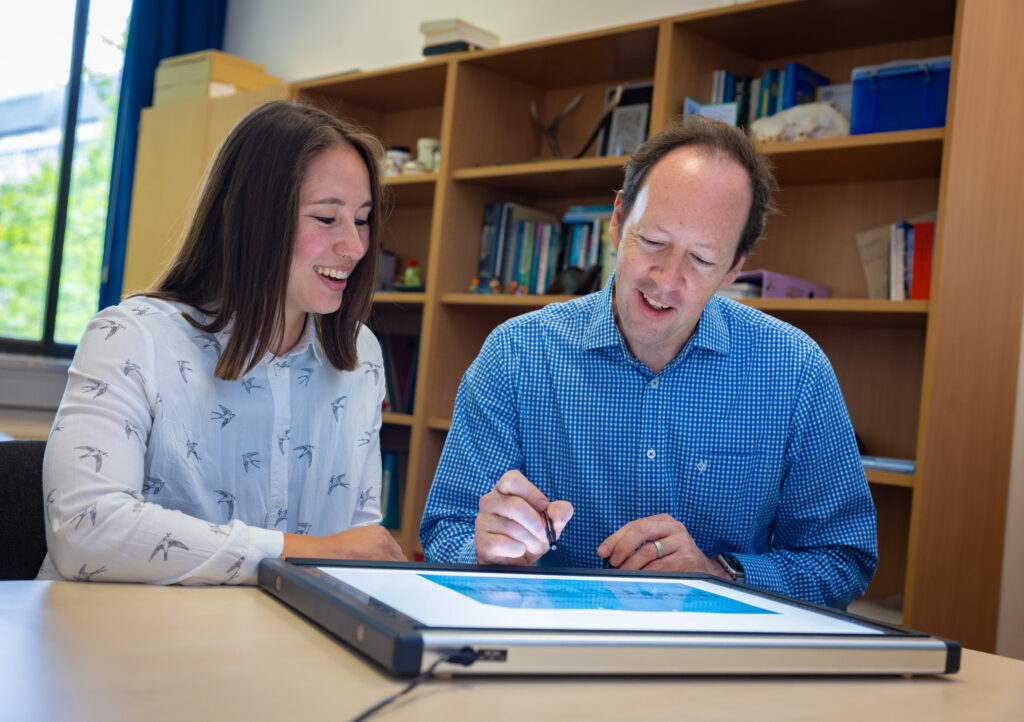 Eine Frau und ein Mann sitzen am Tisch und betrachten Daten auf einer Anzeigetafel, die auf dem Tisch liegt. Im Hintergrund sind Bücherregale.