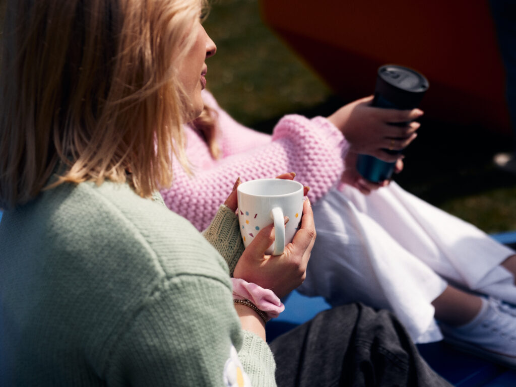 Im Vordergrund eine Person, deren Profil nur leicht erkennbar ist, und die einen Kaffeebecher hält. Im Hintergrund eine weitere Person mit Tumbler. Beide sitzen.