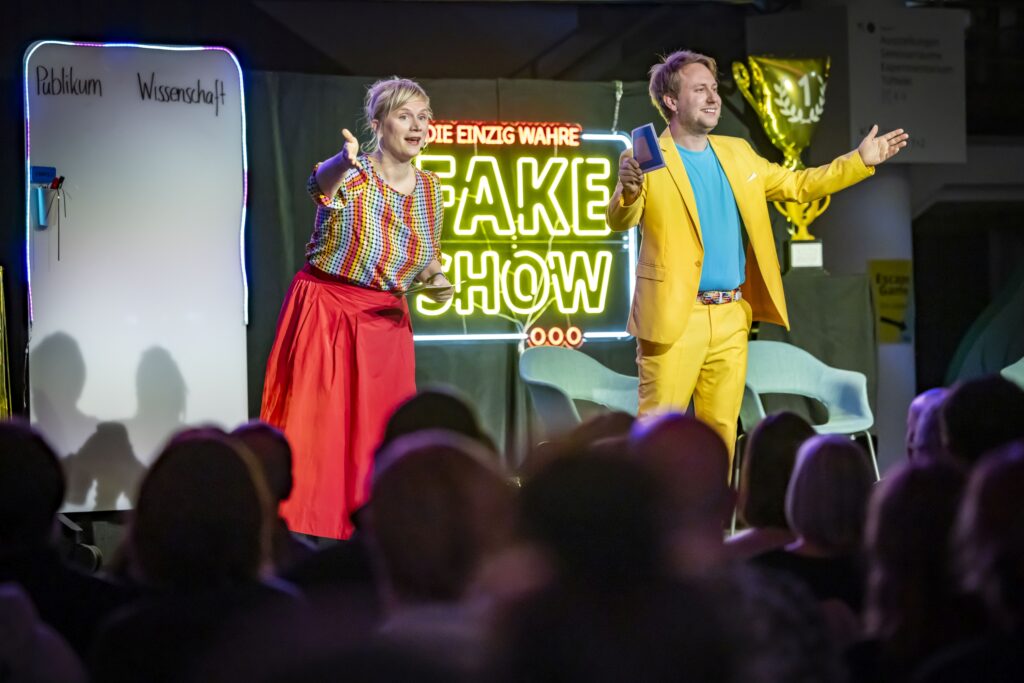 Eine Frau und ein Mann stehen mit einladend ausgestreckten Armen auf der Bühne. Unten im Bild unscharf Menschen aus dem Publikum.