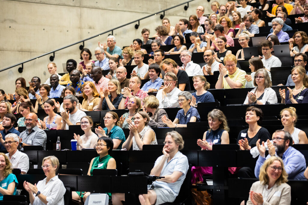 Teilnehmer*innen sitzen in Hörsal und applaudieren.