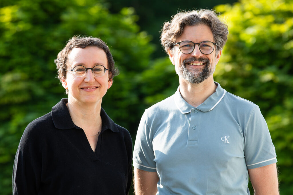 Eine FRau in schwarzer Bluse und ein Mann in grauem T-Shirt stehen vor einem belaubten Hintergrund in der Sonne.