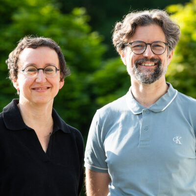 Eine FRau in schwarzer Bluse und ein Mann in grauem T-Shirt stehen vor einem belaubten Hintergrund in der Sonne.