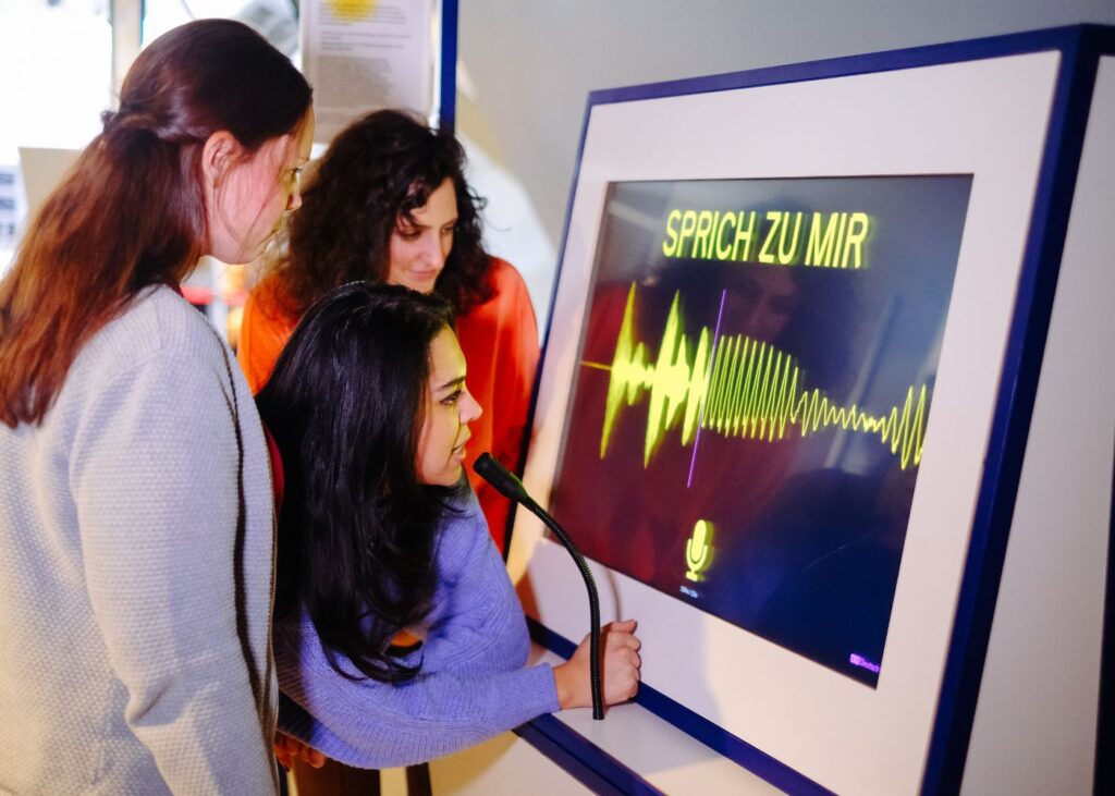 Drei Frauen stehen vor einem Bildschirm, auf dem ' Sprich mit mir' steht, und testen das Gerät