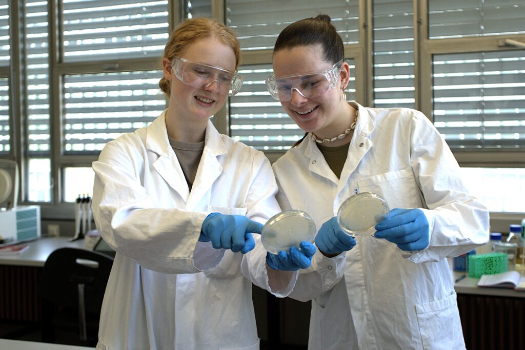 Marielle Rieks (links) und Hannah Bunte (rechts) halten im Labor je eine Petrischale und schauen gemeinsam auf eine der Petrischalenn
