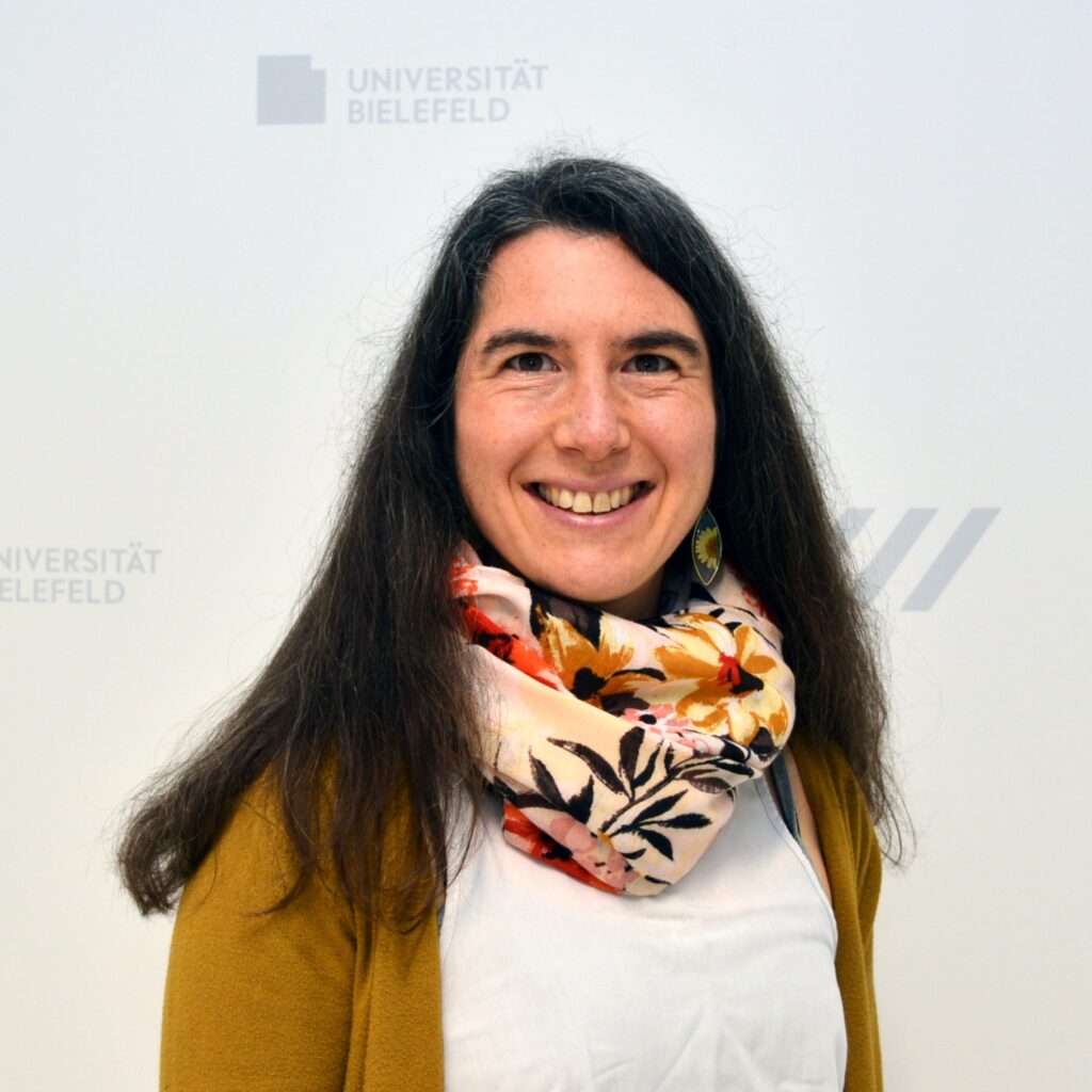 Bild einer dunkelhaarigen Frau vor einem Uni-Hintergrund.