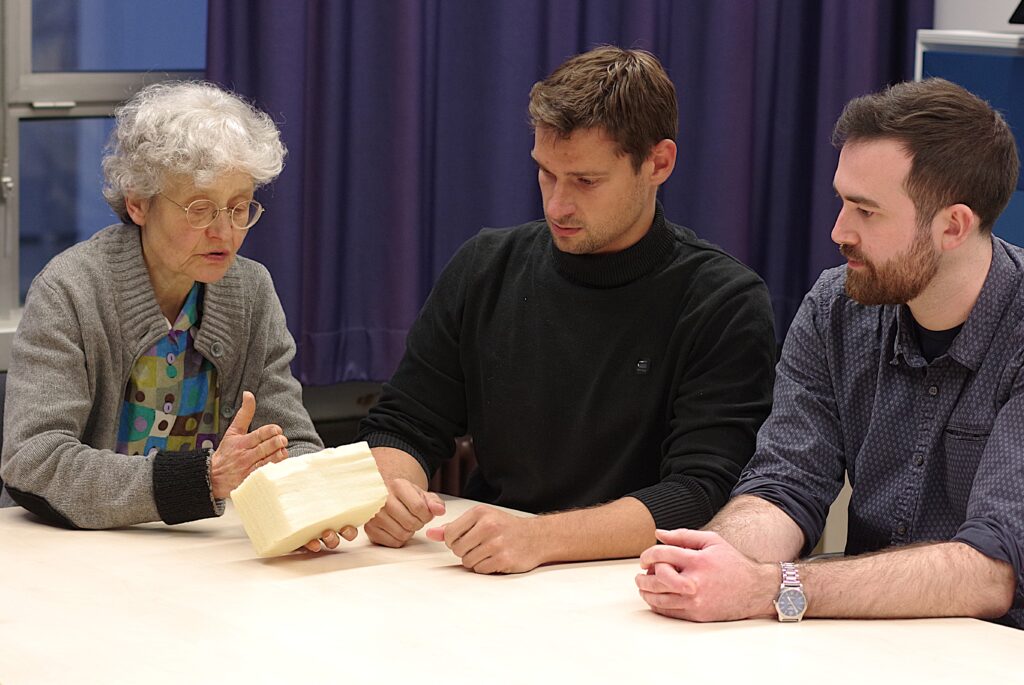 Drei Personen an einem Tisch. Eine Frau hält ein Stück Schaumstoff, die Männer neben ihr schauen darauf.