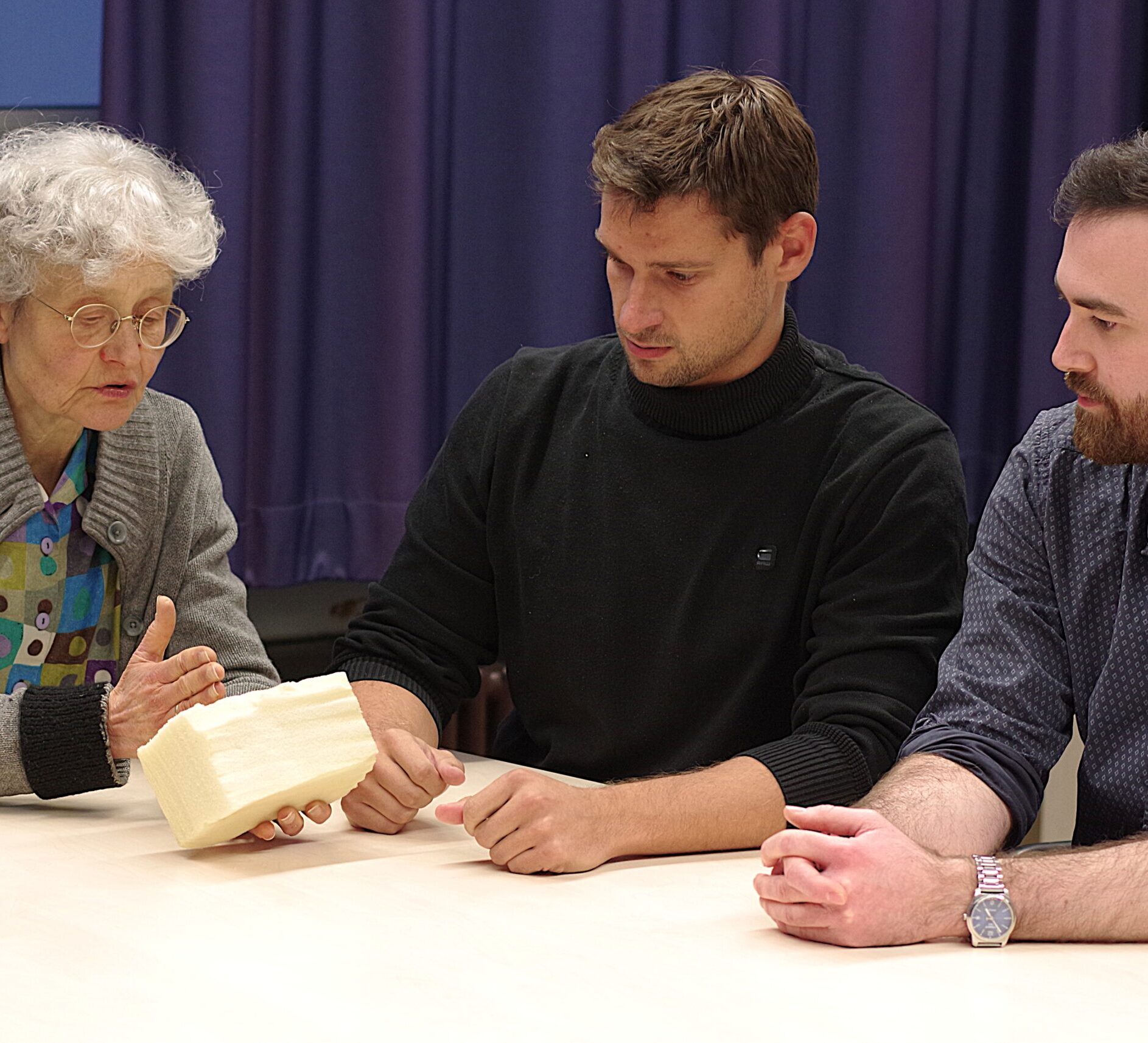 Three people at a table. A woman is holding a piece of foam, and the men next to her are looking at it.