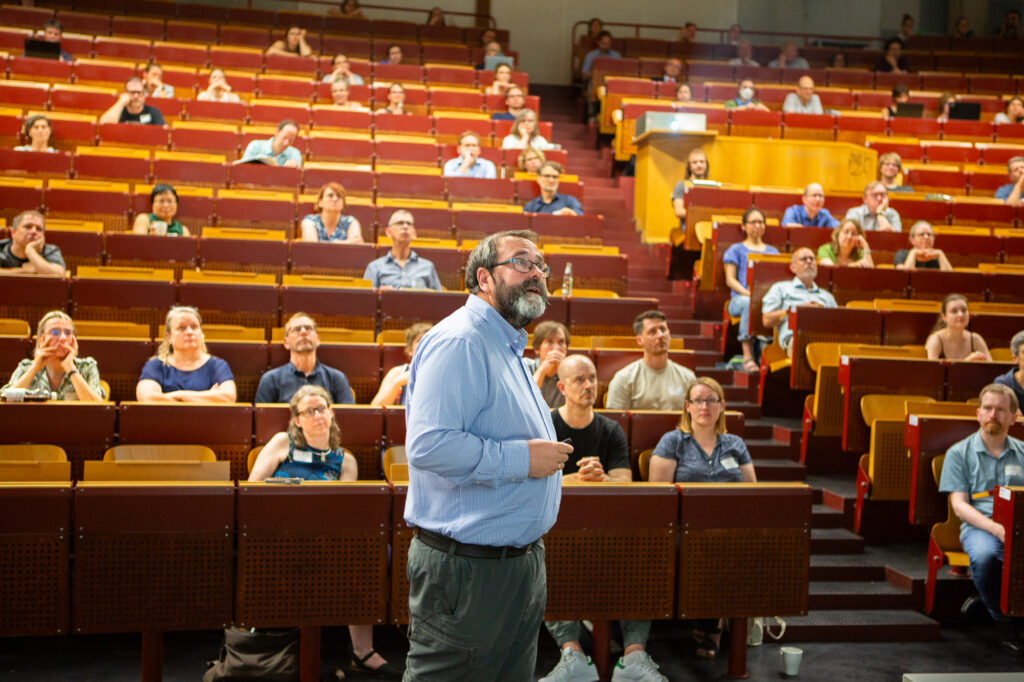 Ein Vortragender steht vorne in einem großen Hörsaal und spricht zu zahlreichen Zuhörerinnen und Zuhörern, die in mehreren Reihen sitzen und aufmerksam zuhören.