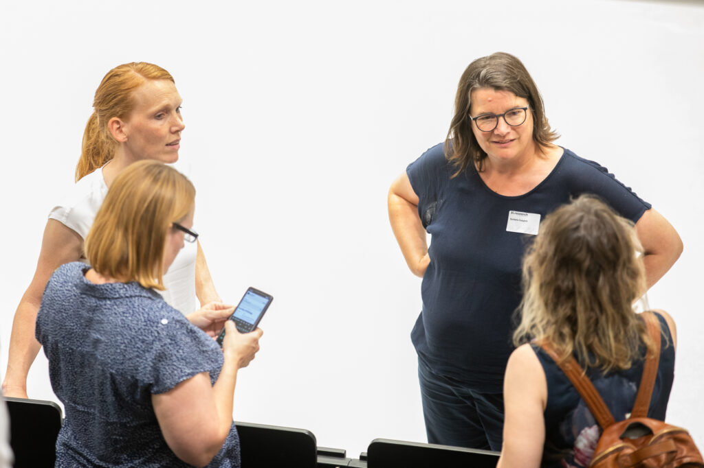 Vier Personen stehen in einem hellen Raum zusammen und unterhalten sich; eine Person schaut auf ihr Smartphone, während die anderen im Gespräch sind.