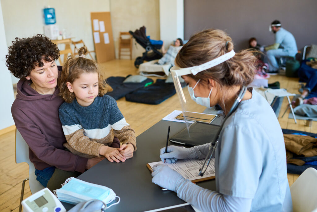 Junge Ärztin füllt medizinisches Formular vor einer Frau aus, deren kleiner Sohn neben dem Schreibtisch sitzt und sich über Unwohlsein beklagt.