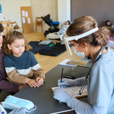 Junge Ärztin füllt medizinisches Formular vor einer Frau aus, deren kleiner Sohn neben dem Schreibtisch sitzt und sich über Unwohlsein beklagt.