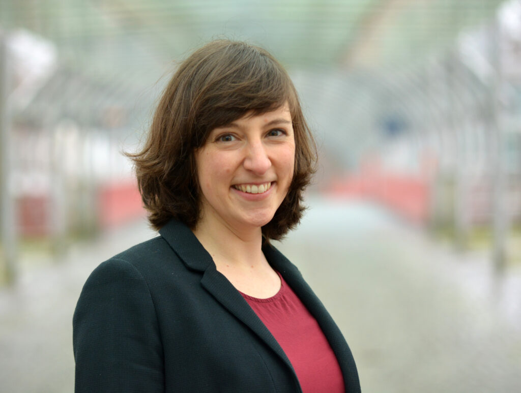 Bild einer Frau mit braunen Haaren in einem schwarzen Blazer und rotem T-Shirt vor verschwommenem Hintergrund.