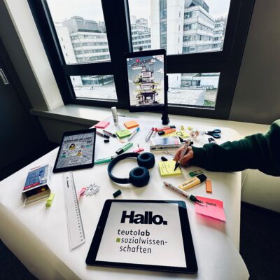 Schreibtisch in einem Büro mit Blick auf moderne Gebäude durch große Fenster. Auf dem Tisch liegen Tablets, Kopfhörer, Marker, Haftnotizen und weiteres Arbeitsmaterial; eine Person schreibt auf einen gelben Notizzettel. Auf einem Tablet steht „Hallo. teutolab sozialwissenschaften“.