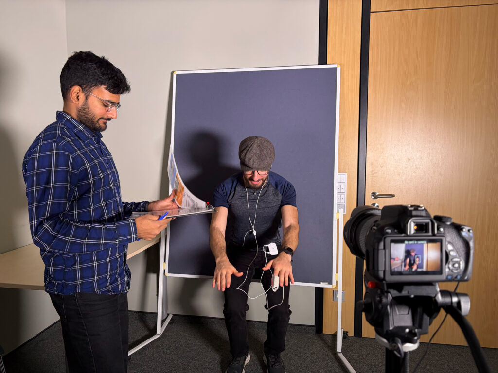 Ein Mann in kartiertem Hemd steht mit einem Bogen Papier in der Hand. Ein anderer Mann mit Mütze sitzt vor einer blauen Leinwand. Im Vordergrund eine Kamera.
