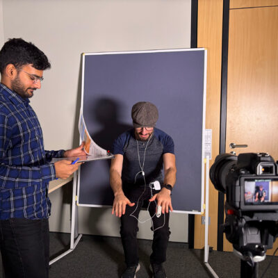 Ein Mann in kartiertem Hemd steht mit einem Bogen Papier in der Hand. Ein anderer Mann mit Mütze sitzt vor einer blauen Leinwand. Im Vordergrund eine Kamera.