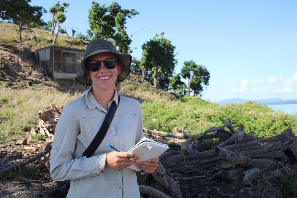Bild einer Frau mit Hut und Sonnenbrille. In der Hand hält sie einen Schreibblock. Im Hintergrund sieht man Affen.