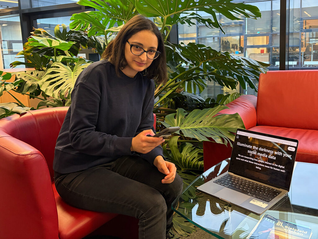 Eine Person mit Brille sitzt auf einem roten Sofa in einem modernen Innenraum mit großen Fenstern und vielen grünen Pflanzen. Sie hält ein Smartphone in der Hand. Auf einem Glastisch steht ein aufgeklappter Laptop mit einer Website über Social-Media-Daten.