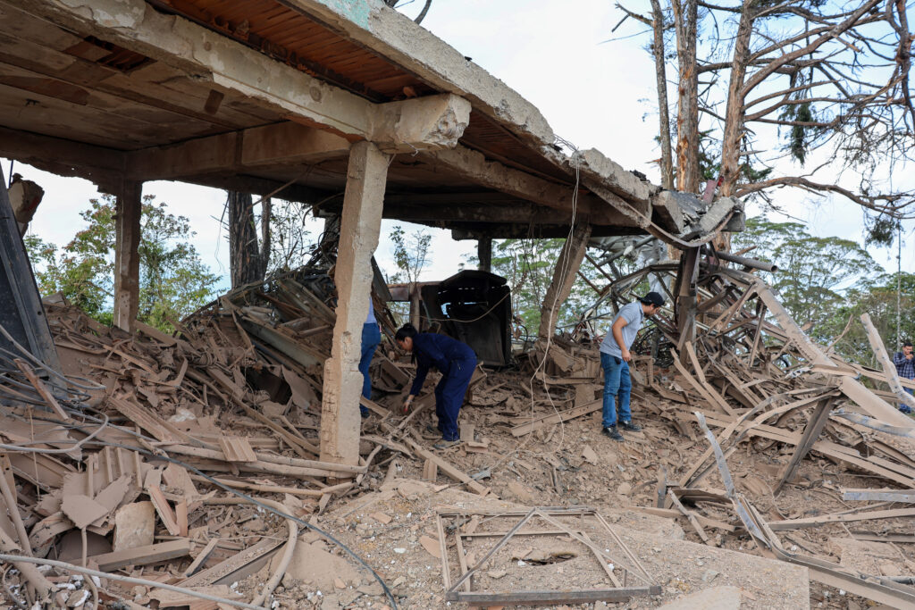 Menschen durchsuchen die Trümmer eines stark beschädigten Betongebäudes; verbogene Metallteile und Schutt liegen unter einer teilweise eingestürzten Decke, im Hintergrund stehen Bäume.