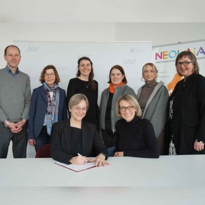 Gruppen von Menschen vor einem Aufsteller mit der Aufschrift NEOLAiA. Im Vordergrund unterschreibt Rektorin Angelika Epple ein Dokument.
