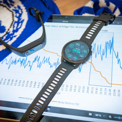 Eine Smartwatch auf einem Holztisch zeigt Gesundheitsdaten an, daneben liegen ein Fitnessarmband und ein blauer Schal. Auf einem Tablet im Hintergrund ist eine Grafik mit wellenförmigen Linien zu sehen, die Herzfrequenz und Stresslevel abbilden.