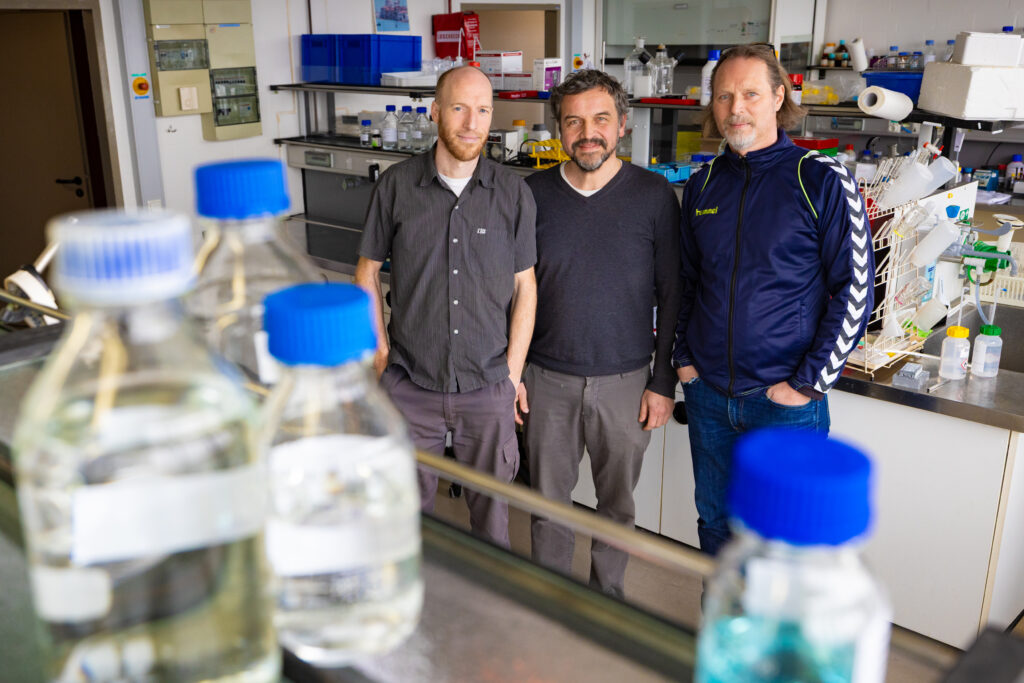 Drei männliche Personen in einer Laborumgebung blicken in die Kamera. Im Vordergund sieht man Plastikfläschchen.