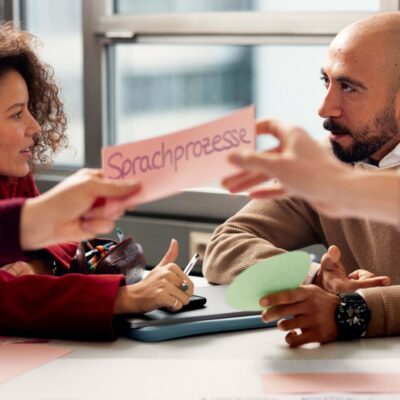 Zwei Personen sitzen an einem Tisch und diskutieren. Davor halten zwei Arme eine Karte mit der Aufschrift „Sprachprozesse“ über einem ausgebreiteten Flipchart-Poster mit weiteren beschrifteten Karten und Notizen.