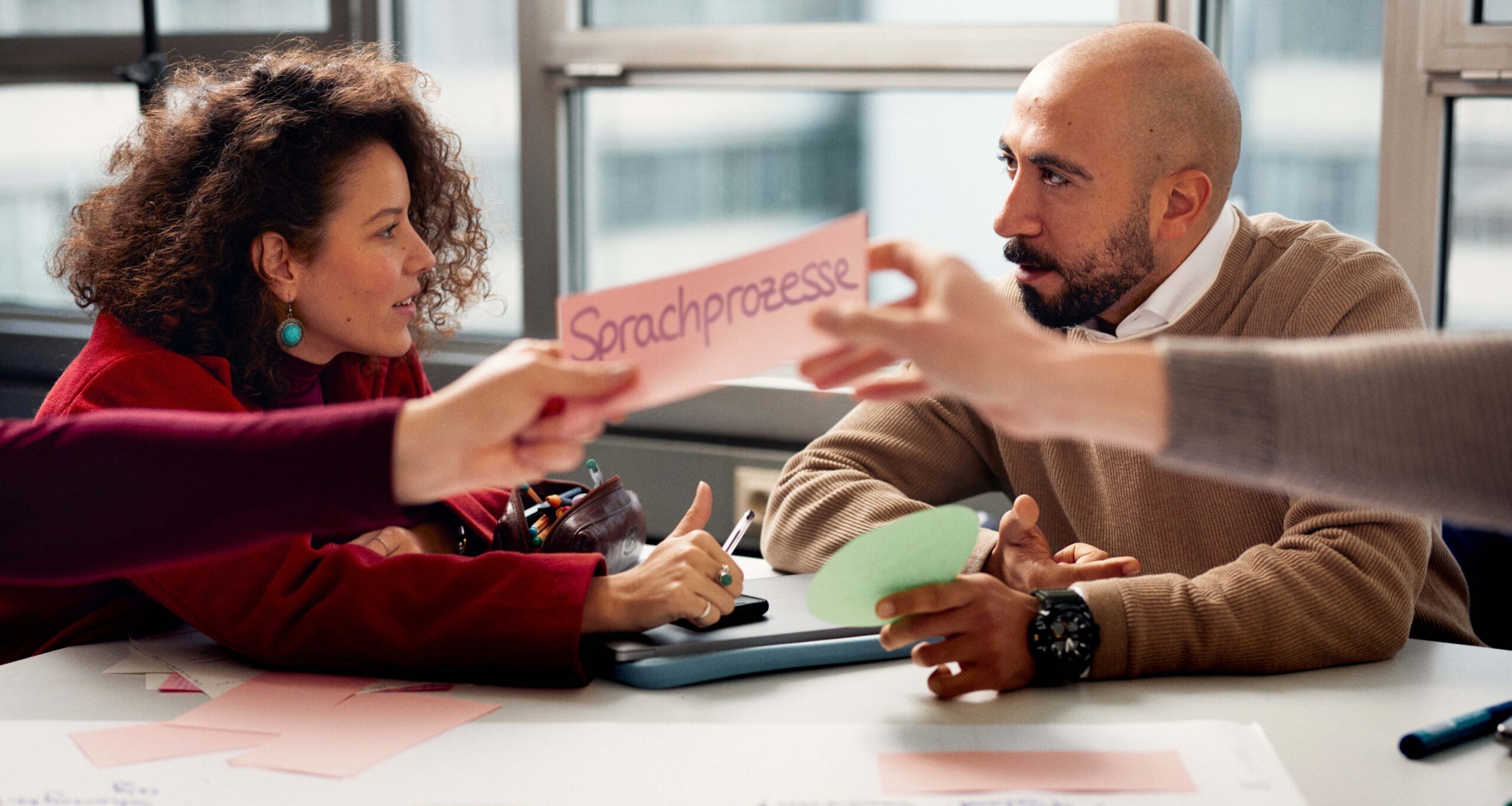 Zwei Personen sitzen an einem Tisch und diskutieren. Davor halten zwei Arme eine Karte mit der Aufschrift „Sprachprozesse“ über einem ausgebreiteten Flipchart-Poster mit weiteren beschrifteten Karten und Notizen.