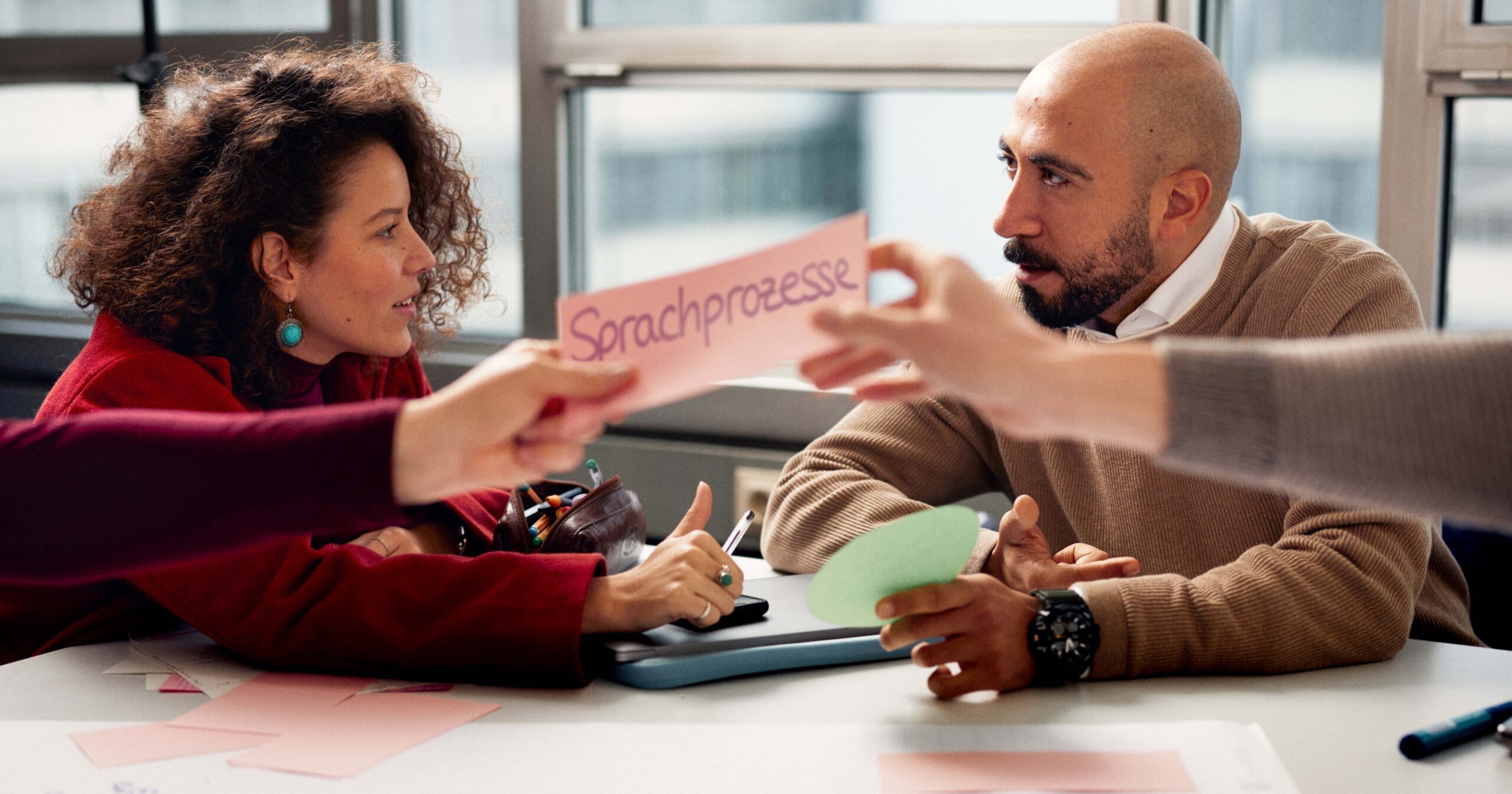 Two people sit at a table discussing. In front of them, two arms hold a card labelled “Sprachprozesse” above a spread-out flipchart poster with additional labelled cards and notes.